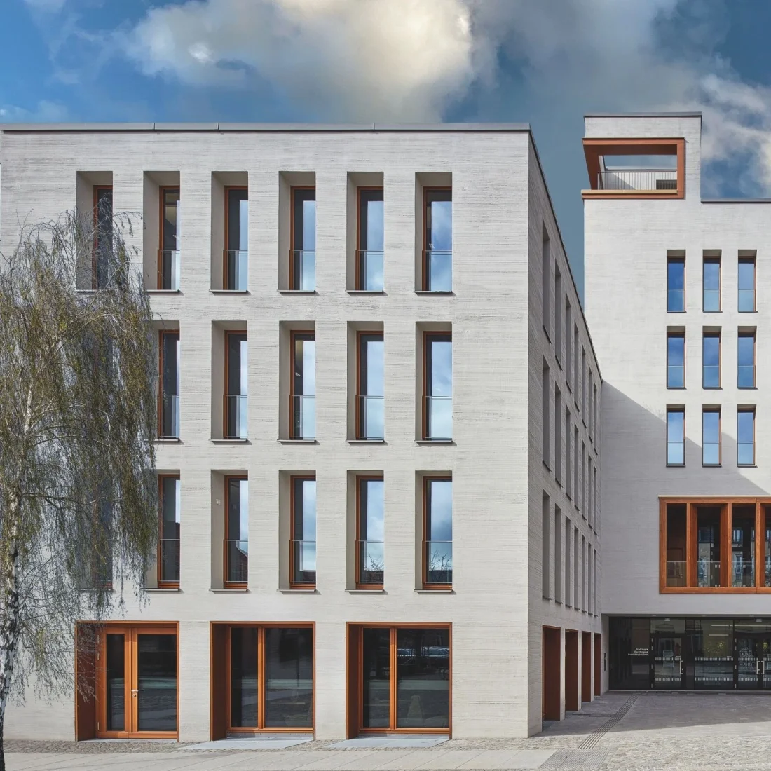 Das moderne Rathaus in Bernau zeichnet sich durch eine klare, strukturierte Fassade aus, die in hellem, sandfarbenem Stein gestaltet ist. Schmale, hohe Fenster gliedern die Fassade rhythmisch und werden durch die dezente Glasabsturzsicherung VITRUM von Abel ergänzt, die sich nahtlos in die Architektur einfügt. Die warmen Holzrahmen der Fenster setzen einen eleganten Kontrast zur Steinverkleidung und verleihen dem Gebäude eine moderne, einladende Note.