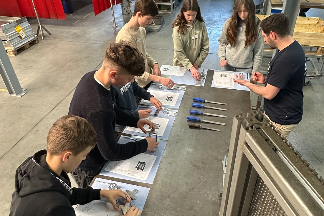 Das Bild zeigt eine Gruppe junger Menschen, die an einem Tisch in einer industriellen Werkstatt oder Ausbildungsstätte arbeiten. Die Teilnehmer, bestehend aus Jungen und Mädchen, tragen Freizeitkleidung und sind mit technischen Zeichnungen oder Anleitungen beschäftigt. Vor ihnen auf dem Tisch liegen Werkzeuge wie Schraubendreher sowie Metallteile, die sie anscheinend zusammenbauen oder untersuchen.  Ein erwachsener Mann, möglicherweise ein Ausbilder, steht auf der rechten Seite und gibt Anweisungen oder erklärt etwas. Die Umgebung sieht nach einer Produktionshalle oder einer Lehrwerkstatt aus, mit metallischen Strukturen und industriellen Elementen im Hintergrund.  Die Szene deutet darauf hin, dass es sich um eine praktische Lerneinheit handelt – möglicherweise im Rahmen eines Berufsorientierungstags wie dem „Girls’ and Boys’ Day“, an dem Jugendliche verschiedene Berufsfelder erkunden können.