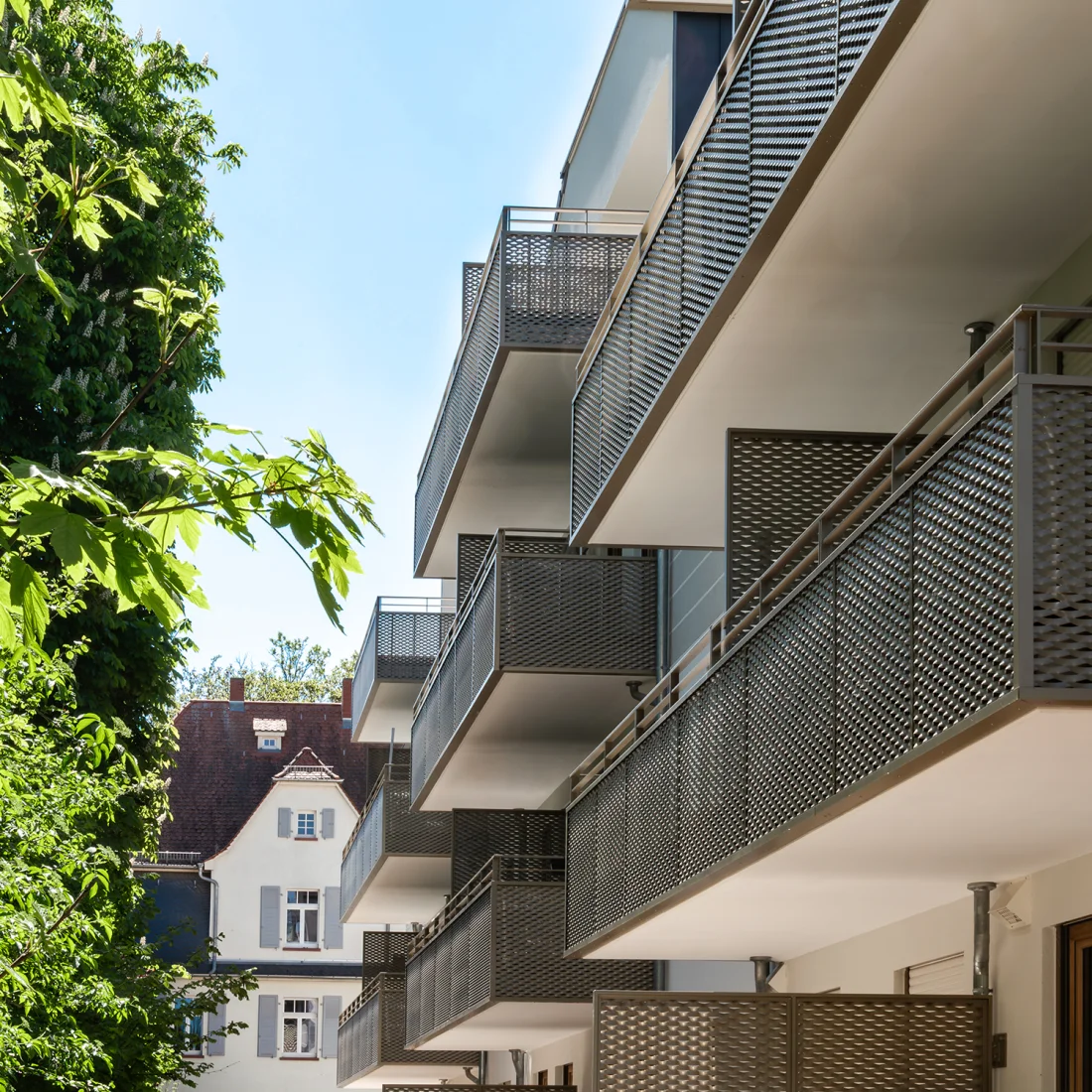 Dieses Bild zeigt die Seitenansicht eines modernen Mehrfamilienhauses mit mehreren Stockwerken, die alle mit Balkonen ausgestattet sind. Die Balkonbrüstungen bestehen aus einem dunklen, perforierten Metallgitter, das ein interessantes Muster aufweist. Die Untersichten der Balkone sind hell gehalten und bilden einen Kontrast zu den dunklen Brüstungen. Links im Vordergrund ist ein großer, grüner Baum mit dichtem Laubwerk zu sehen, der einen natürlichen Rahmen bildet. Im Hintergrund, etwas verschwommen, ist ein älteres Wohnhaus mit hellen Fassaden und dunklem Dach zu erkennen, was auf eine Mischung aus Alt- und Neubau in der Umgebung hindeutet. Der Himmel ist hellblau und klar, was auf gutes Wetter schließen lässt.