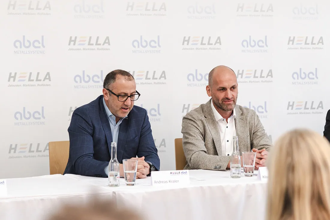 Auf dem Bild sind die Geschäftsführer Andreas Kraler von Hella und Klaus Peter Abel von Abel Metallsysteme während einer Pressekonferenz zu sehen. Die Konferenz dient der Vorstellung der Fusion von zwei Produkten: Sonnenschutzsysteme von Hella und integrierte Absturzsicherungen von Abel Metallsysteme. Beide Geschäftsführer sitzen nebeneinander und präsentieren die neu fusionierten Produkte der beiden Unternehmen.