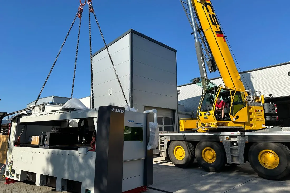 Auf dem Bild hebt ein gelber Autokran einen grauen Laser vor der Produktionshalle von Abel Metallsysteme in die Höhe. Im Hintergrund strahlt der Himmel, was die Szene besonders hervorhebt.