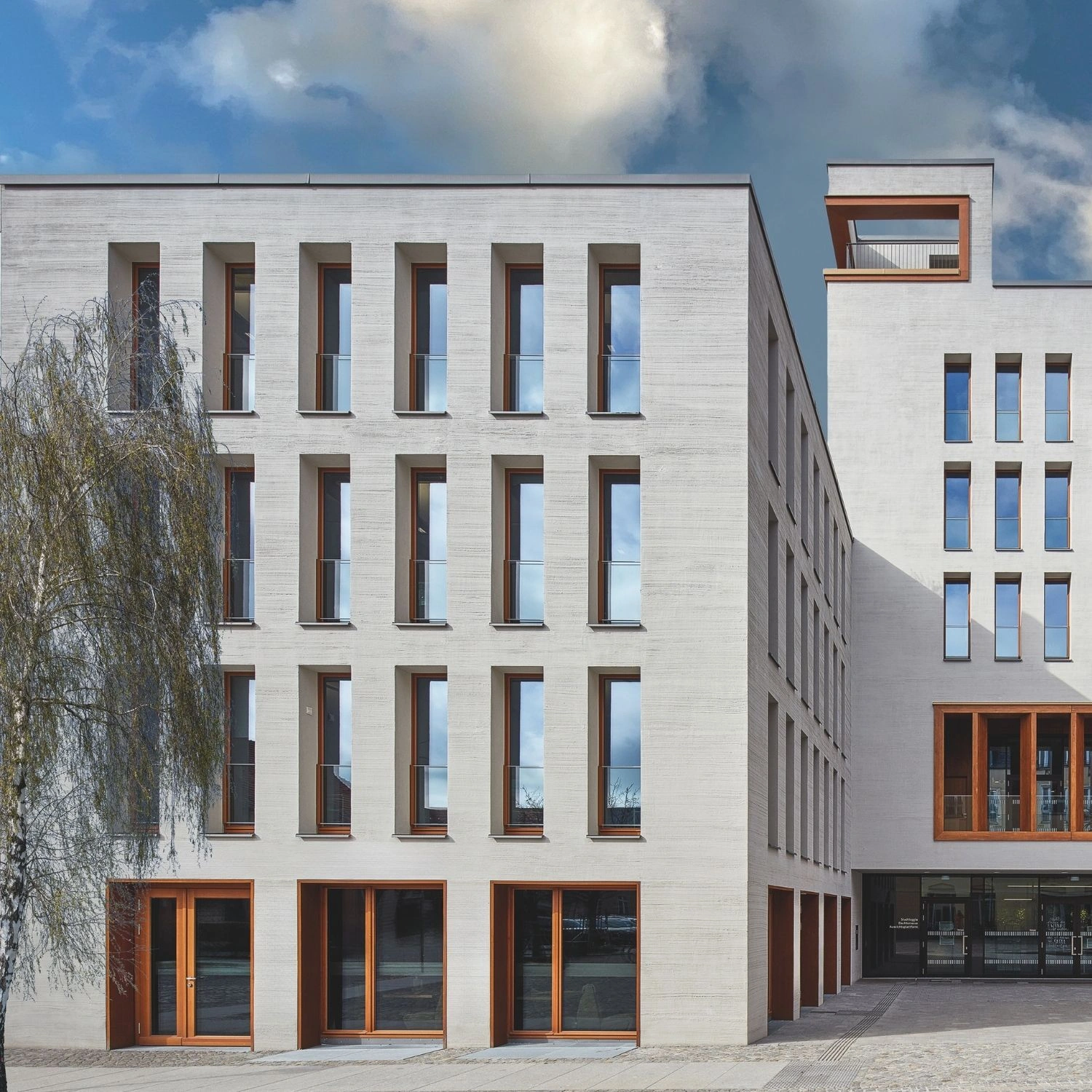 Das moderne Rathaus in Bernau zeichnet sich durch eine klare, strukturierte Fassade aus, die in hellem, sandfarbenem Stein gestaltet ist. Schmale, hohe Fenster gliedern die Fassade rhythmisch und werden durch die dezente Glasabsturzsicherung VITRUM von Abel ergänzt, die sich nahtlos in die Architektur einfügt. Die warmen Holzrahmen der Fenster setzen einen eleganten Kontrast zur Steinverkleidung und verleihen dem Gebäude eine moderne, einladende Note.