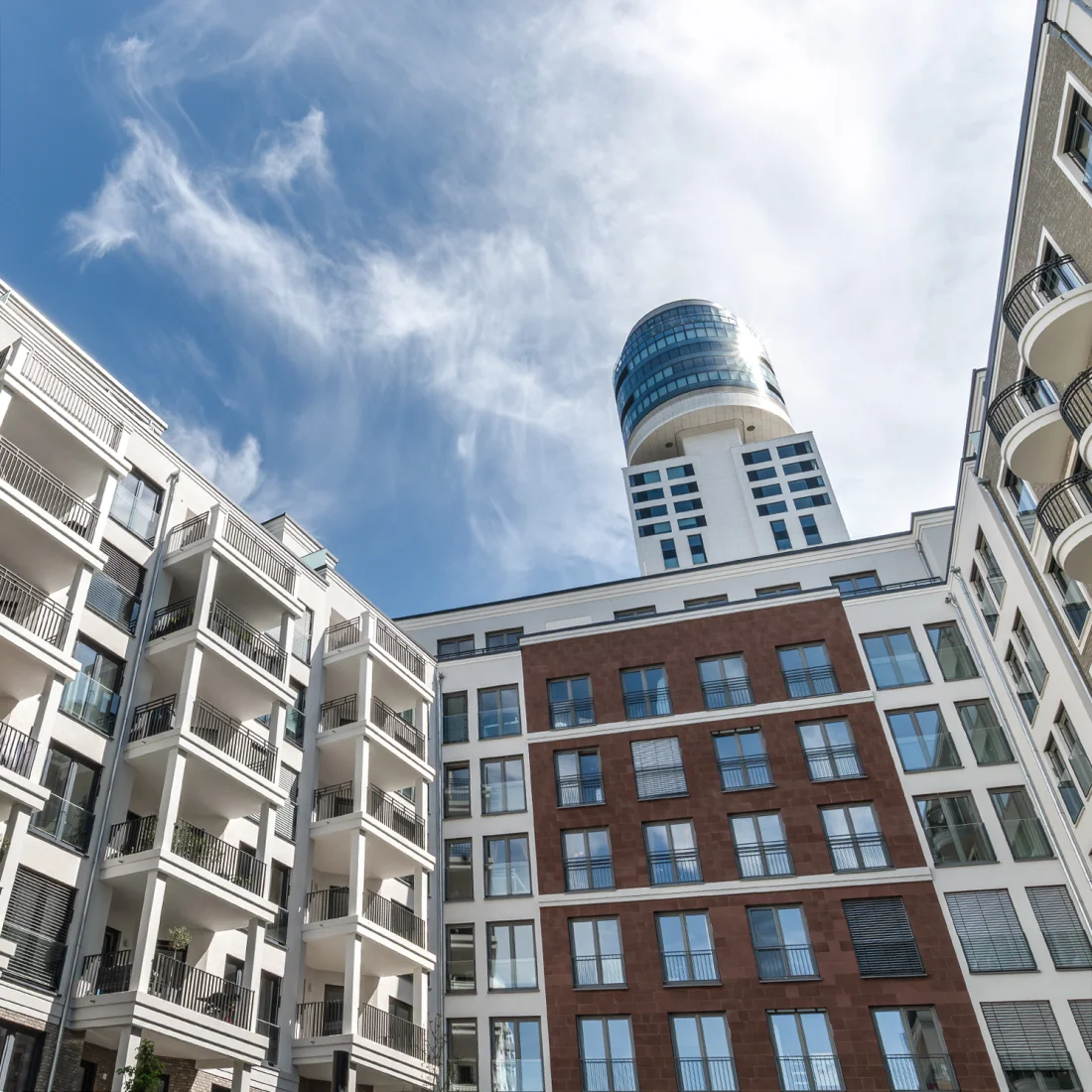 Moderne Architektur trifft auf urbanes Flair: Ein Blick von unten auf eine Gruppe stilvoller Wohngebäude, die sich harmonisch in die Stadtlandschaft einfügen, mit einem markanten Hochhaus im Hintergrund. Der strahlend blaue Himmel mit wenigen Wolken unterstreicht die klare Linienführung und Eleganz der Fassaden.