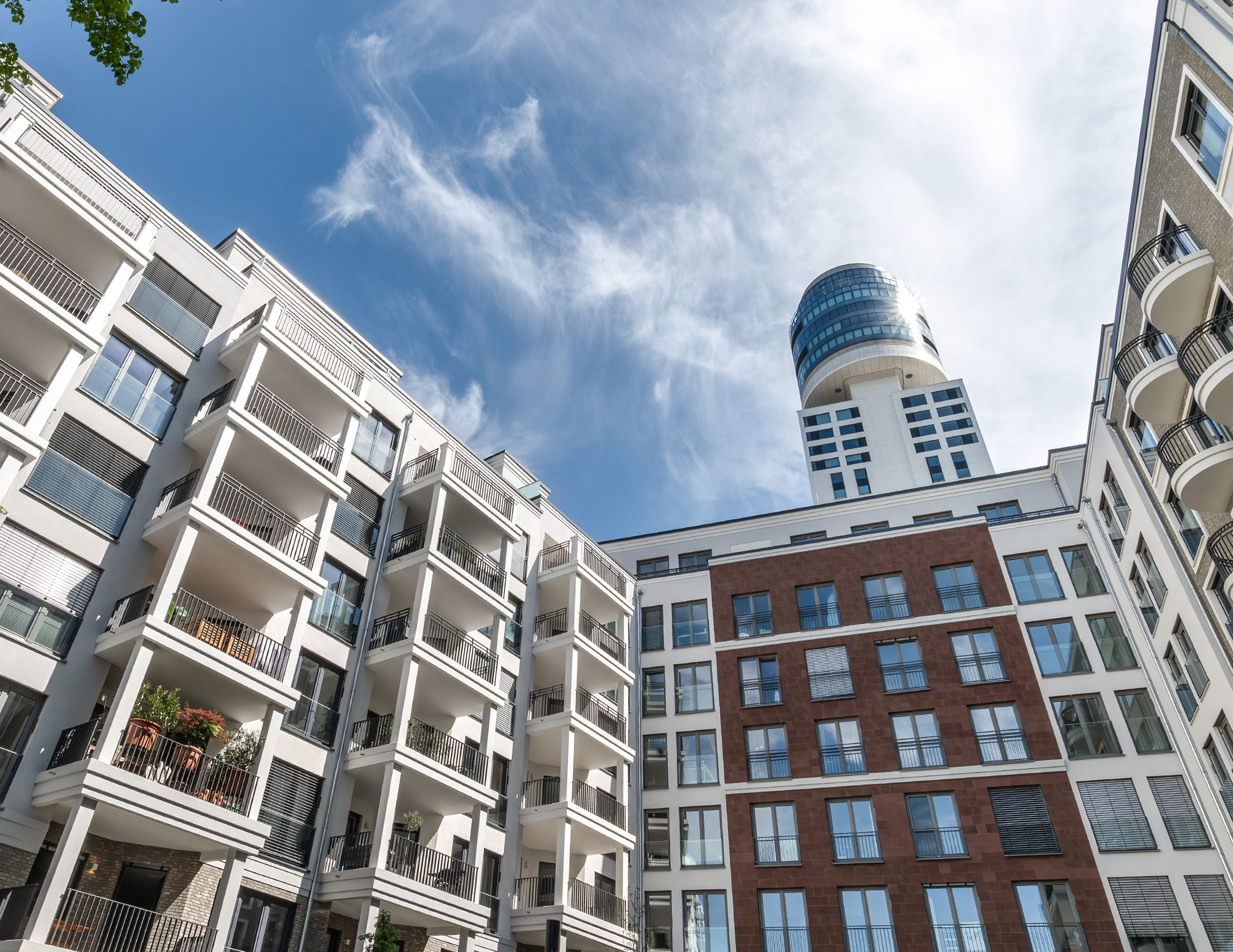 Moderne Architektur trifft auf urbanes Flair: Ein Blick von unten auf eine Gruppe stilvoller Wohngebäude, die sich harmonisch in die Stadtlandschaft einfügen, mit einem markanten Hochhaus im Hintergrund. Der strahlend blaue Himmel mit wenigen Wolken unterstreicht die klare Linienführung und Eleganz der Fassaden.