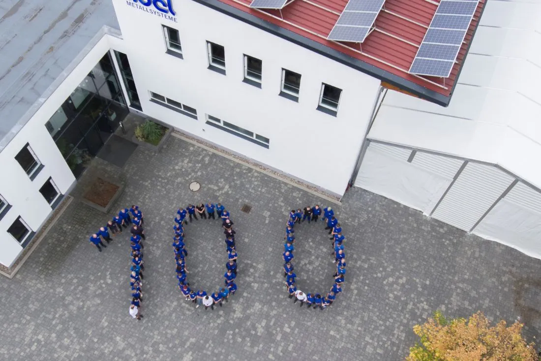 Auf dem Bild ist eine große „100“ zu sehen, die von den Mitarbeitern des Unternehmens Abel Metallsysteme gebildet wird. Die Formation steht vor einem weißen Verwaltungsgebäude und markiert das 100-jährige Bestehen des Unternehmens. Die Aufnahme wurde aus der Luft gemacht, um die beeindruckende Zahl und die Zusammensetzung der Mitarbeiter deutlich zu zeigen.