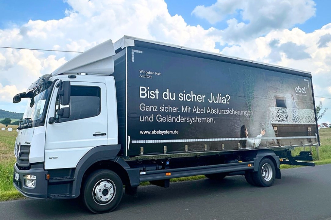 Das Bild zeigt einen modernen LKW, der auf einer Landstraße vor einem weiten Feld steht. Der Himmel ist strahlend blau, durchzogen von leichten Quellwolken, was einen idyllischen Sommertag andeutet. Auf der Seite des LKWs prangt eine neue Plane, die eine kreative Werbebotschaft trägt. Der Text auf der Plane lautet: „Bist du sicher, Julia? Ganz sicher. Mit Abel Absturzsicherungen und Geländersystemen.“ Die Szene auf der Plane ist eine humorvolle Abwandlung der berühmten Balkonszene aus „Romeo und Julia“, wobei Julia hier in einem sicheren Geländersystem steht, das Abel bewirbt. Der LKW, die Werbung und die sommerliche Landschaft harmonieren zu einem einprägsamen Bild.