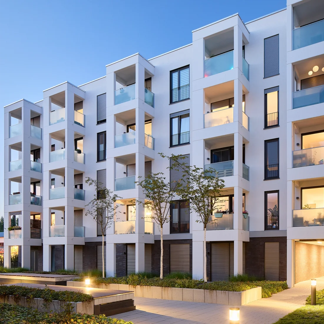     Anfrage erfolgreich  Ein modernes, mehrstöckiges Wohngebäude mit weißer Fassade und vielen Balkonen ist zu sehen. Die Balkone haben Glasbrüstungen und einige sind beleuchtet, was auf bewohnte Einheiten hindeutet. Die Fenster sind überwiegend dunkel oder zeigen ebenfalls Innenbeleuchtung. Im unteren Bereich des Gebäudes ist eine Mischung aus weißem Putz und dunkleren, möglicherweise gemauerten Elementen erkennbar. Vor dem Gebäude befinden sich kleine Bäume und eine gepflasterte Fläche, die von unten beleuchtet wird, was eine warme Atmosphäre schafft. Die Beleuchtung im Inneren des Gebäudes sowie die äußere Beleuchtung lassen vermuten, dass es sich um Abendstunden handelt.