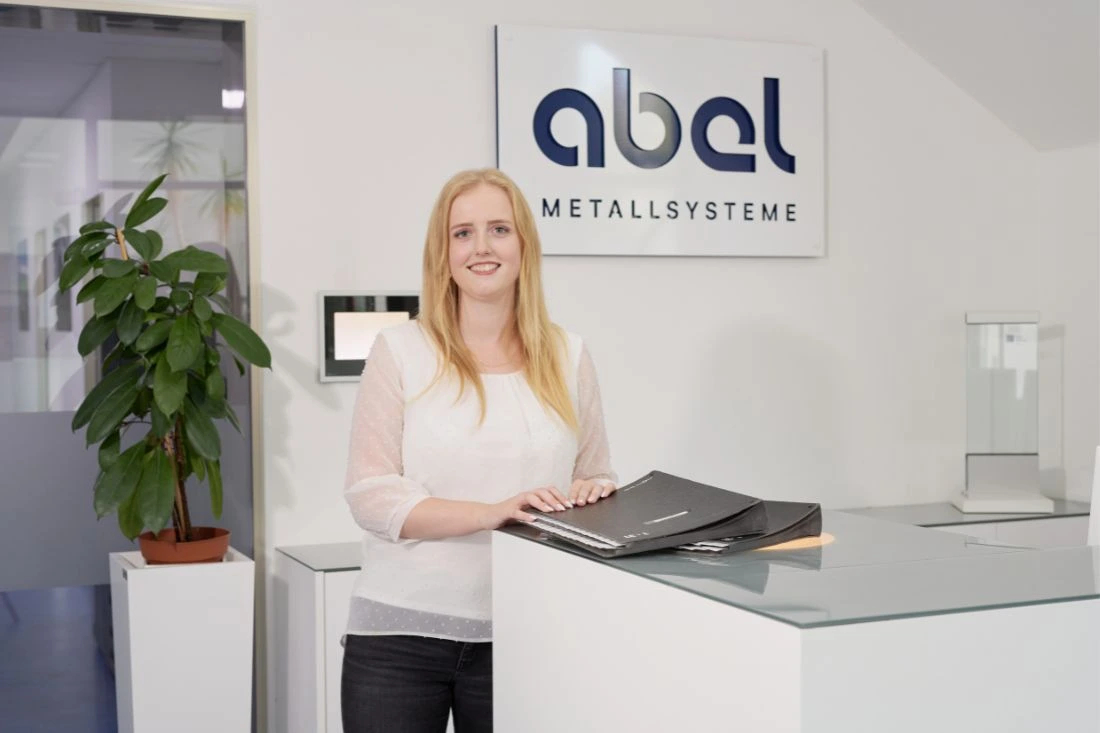 Auf dem Bild ist eine junge Frau in einem modernen Büro zu sehen. Sie sitzt an einem Schreibtisch und arbeitet an einem Computer. Die Frau hat blonde, wellige Haare, die über ihre Schultern fallen. Sie trägt ein lila Oberteil und eine schwarze Jacke. Vor ihr steht ein großer weißer Monitor, und sie lächelt freundlich in die Kamera. Der Stuhl, auf dem sie sitzt, ist grün, und der Raum wirkt hell und professionell eingerichtet. Die Atmosphäre im Bild ist angenehm und zeigt eine konzentrierte, aber freundliche Arbeitssituation.