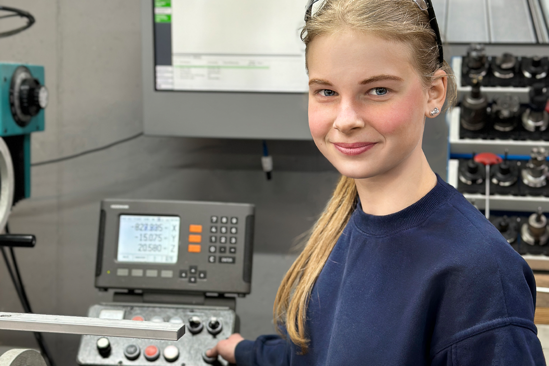 Auf dem Bild ist eine junge Frau mit blonden Haaren, die zu einem Zopf zusammengebunden sind. Auf ihrem Kopf hat sie eine Schutzbrille leicht in die Haare gesteckt sitzen. Sie trägt einen dunkelblauen Abel Metallsysteme Pullover. Sie steht an einer Drehmaschine uns betätigt einen Knopf. Der Fokus liegt auf dem freundlich lächelnden Gesicht der jungen Frau, die als Produktionsmitarbeiterin bei Abelmetallsysteme in der Produktion tätig ist.  